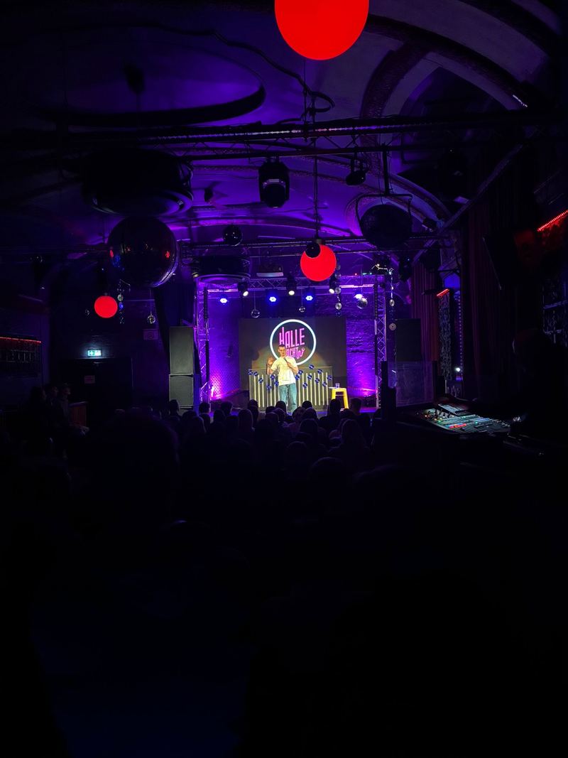 Comedian mit Mikrofon auf kleiner Bühne, Neonschriftzug „Halle lacht“ im Hintergrund