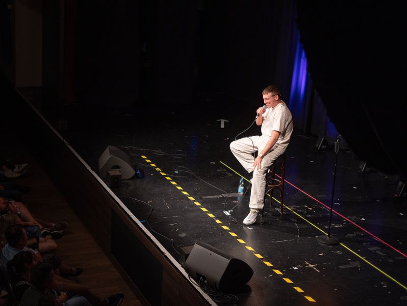 Comedian im weißen Anzug auf einem Barhocker, weite dunkle Bühne von oben
